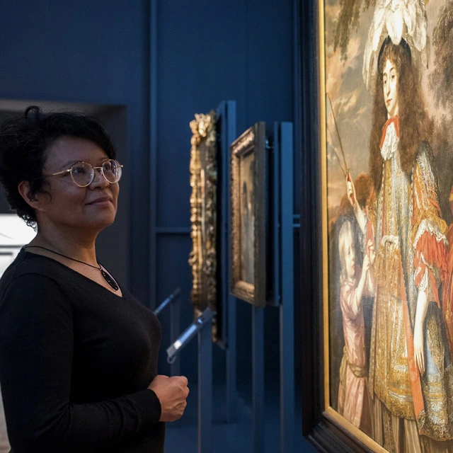 Close-up of a woman gazing at a large colonial painting depicting a noblewoman with children. The woman stands in a dark gallery lit by focused spotlights on the artworks.