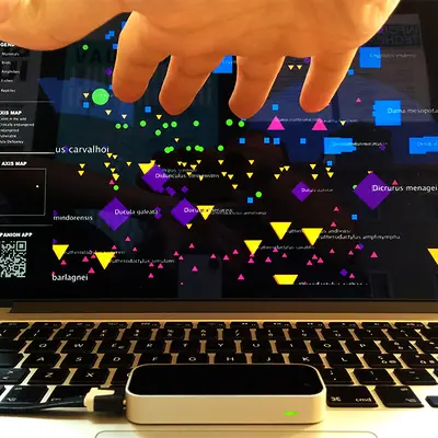Close-up of a hand using Leap Motion gesture control above a laptop keyboard, navigating the 3D extinction visualisation on screen, with species names and coloured shapes visible`