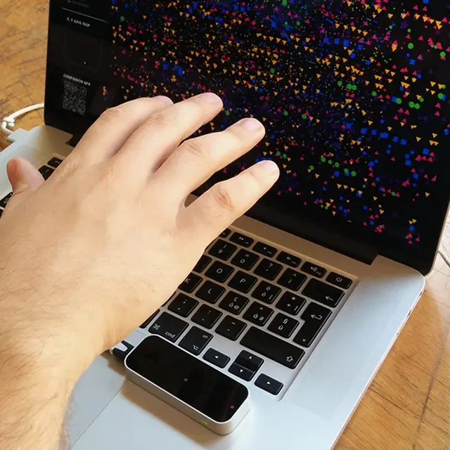 A person’s hand resting on top of a Leap Motion device near a MacBook trackpad while the screen shows the colourful extinction data visualisation with geometric shapes representing animal species.
